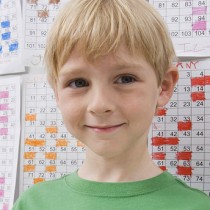 young blonde boy with half smile in elementary school math classroom standing in front of completed worksheets